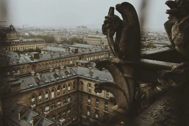 A historic gargoyle looks over a foggy city. Repetitive, brick-built structures line the streets. 