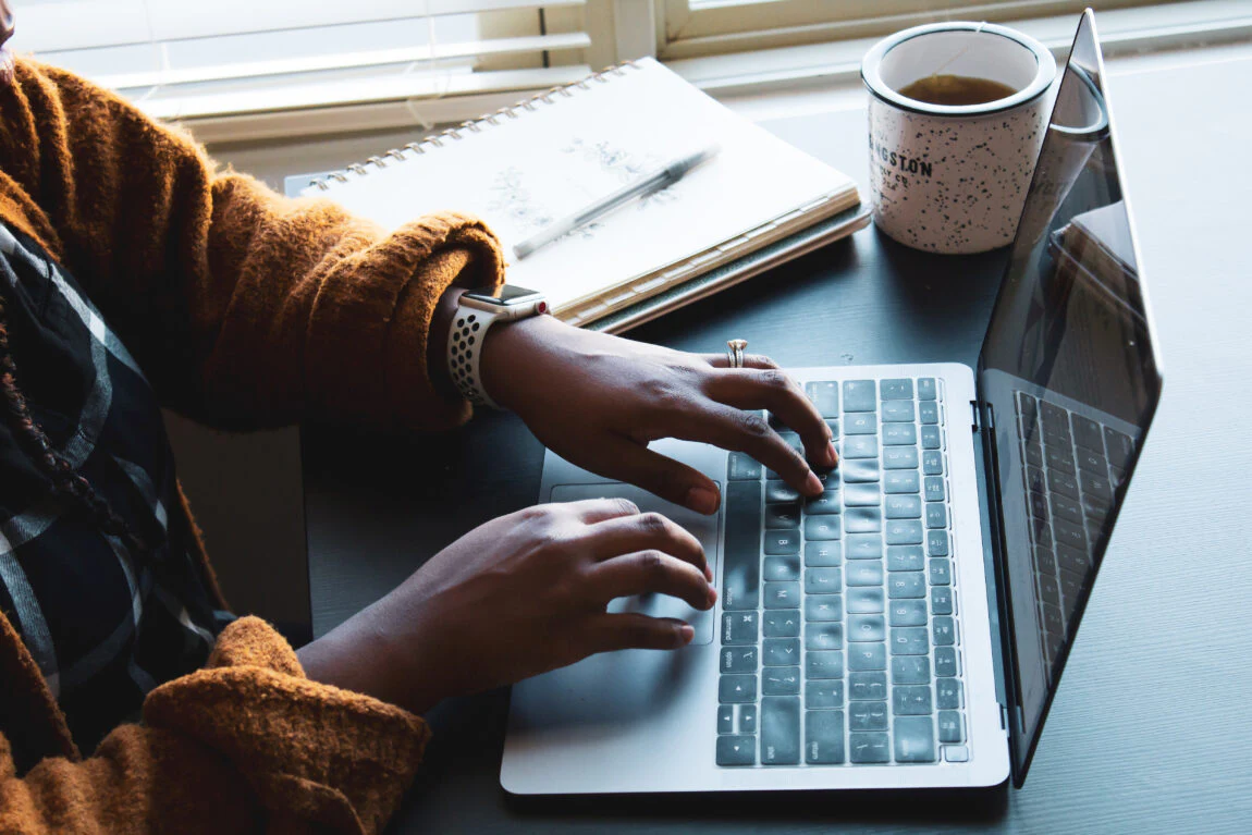 Person wearing a brown sweater typing on a laptop, with a notebook and a mug of coffee placed on a desk near a window.