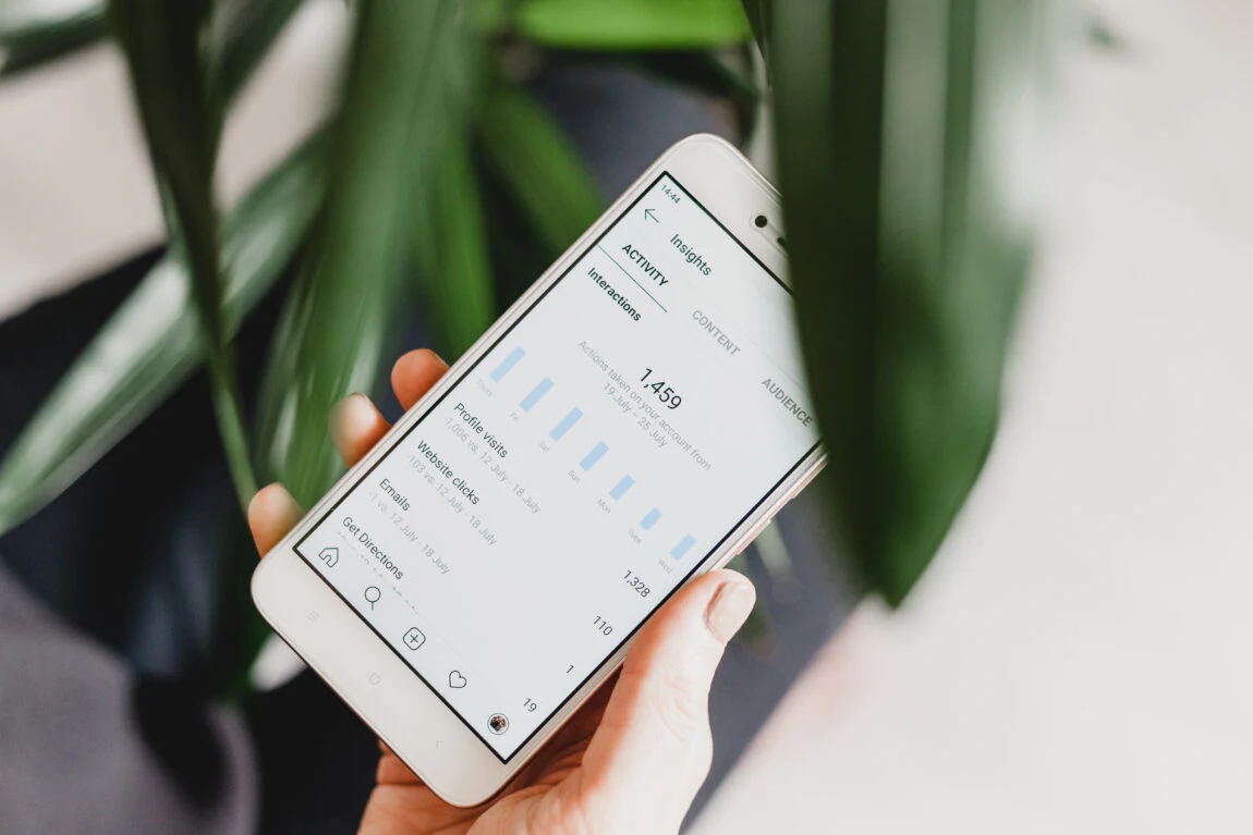 Close-up of a hand holding a smartphone displaying Instagram Insights, showing analytics data such as impressions, profile visits, and interactions against a blurred background with green leaves.
