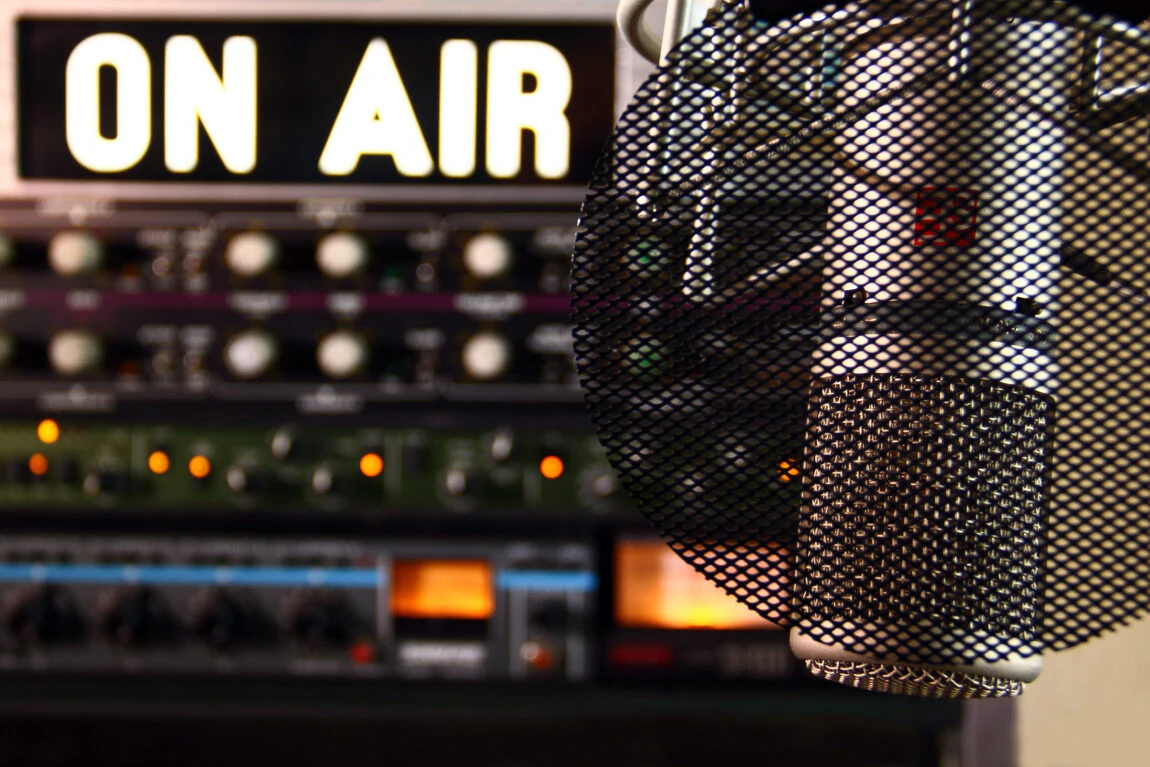 Close-up of a professional microphone with a pop filter in front of a glowing 'ON AIR' sign and audio equipment in a radio studio.