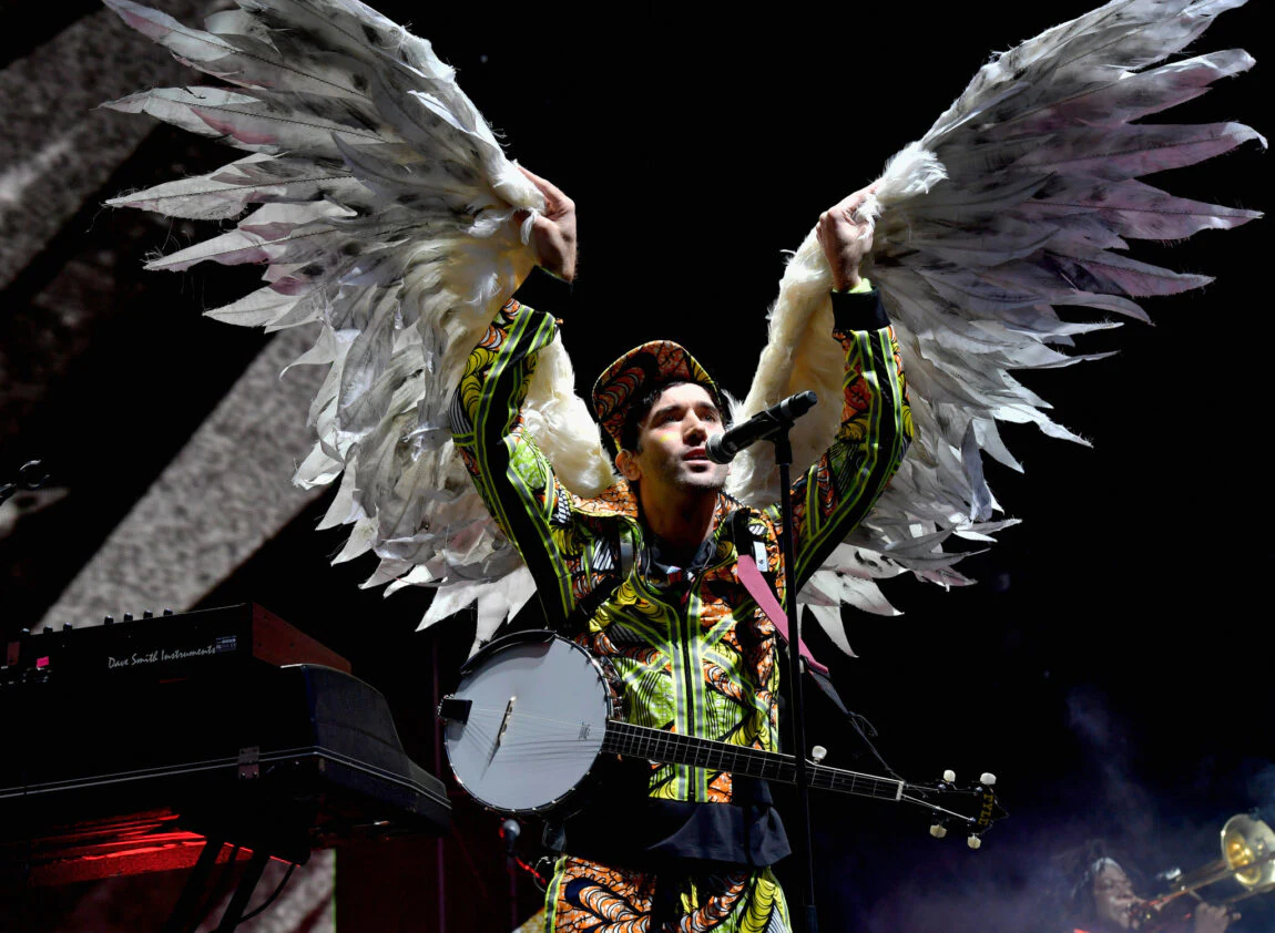 Sufjan Stevens on stage wearing a tracksuit and angel wings. His arms are extended upwards as he sings.